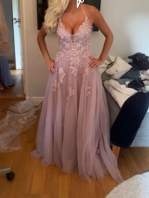 Ljusrosa långklänning med spetsdetaljer - Drömmig ljusrosa långklänning med blommiga spetsapplikationer och tunn tyllkjol. Djup v-ringning, smala axelband och öppen rygg med snörning. Perfekt för bal eller student. Materialet känns lätt och flowy, och detaljerna ger en romantisk vibe. Är 162cm och funkade perfekt med ballerina skor och klackar 