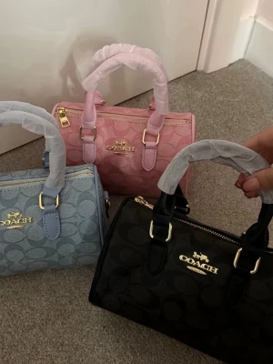 Coach handväska i blått, rosa eller svart - Coach handväska i klassisk barrel-modell med guldfärgade detaljer och dragkedja. Finns i ljusblått, ljusrosa eller svart med Coachs ikoniska mönster. Väskan är tillverkad i slitstark polyester med skinnliknande detaljer och har dubbla handtag.