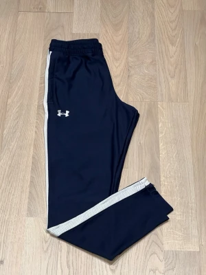 Träningsbyxor från Under Armour med dragkedja - Vit rand längs sidorna och logga pả högra låret. Mjukt material för extra komfort.