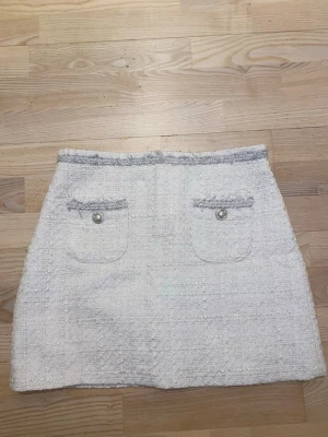 Vit kortkjol från Zara Woman - Vit kortkjol från Zara Woman i strukturerat tyg med två framfickor dekorerade med pärlknappar. Kjolen har diskret dragkedja i bak och glittriga detaljer vid midjan och fickorna.