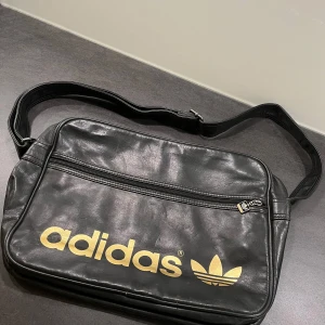 Svart axelväska från Adidas - Klassisk svart axelväska från Adidas med justerbar rem och stort guldfärgat Adidas-tryck på framsidan. Väskan har ett ytterfack med dragkedja och är tillverkad i syntetmaterial som liknar skinn. Perfekt för att bära med sig det viktigaste till skolan eller stan.