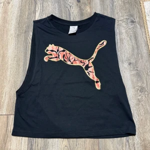 Svart croppad linnetopp Puma Kenza - Säljer en svart croppad linnetopp från Puma x Kenza med ett stort färgglatt Puma-tryck framtill. Toppen är ärmlös med breda axelband och har en avslappnad passform. Perfekt till sommarens alla looks!