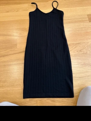 Svart ribbad kortklänning med smala axelband - En svart ribbad kortklänning med smala axelband och v-ringad hals. Klänningen har en enkel och stilren design som är lätt att matcha. Materialet känns  följsamt, perfekt för en avslappnad look.
