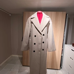 Beige ullkappa från Lindex Extended - Säljer en lång, beige ullkappa från Lindex med dubbelknäppning och stora svarta knappar. Kappan har bred krage, bälte i midjan och raka, långa ärmar. 52 % ull,44% polyester, 2% polyamid, 2 % acryl. Halv foder i satin. 