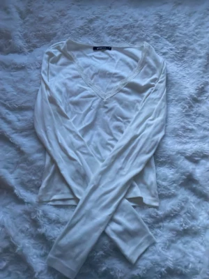 Vit långärmad topp från Ginatricot - Säljer en enkel och stilren vit långärmad topp från Ginatricot. Toppen har v-ringning med spetsdetalj runt halsen och är gjord i ett mjukt, tunt material som känns skönt mot huden. Perfekt att matcha med jeans eller kjol. Den är skrynklig för den har varit vikt länge i min garderob 