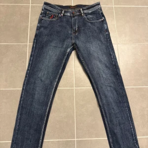 Mörkblå jeans från Kiton, storlek 32 - Snygga mörkblå jeans från Kiton med klassisk femficksdesign och coola röda detaljer på fickor och knapp. Jeansen har en rak passform och är tillverkade i slitstarkt bomullsmaterial med jeansstruktur. Perfekt för dig som gillar stilrena och exklusiva jeans.