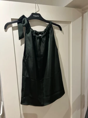 Grön satinlinnetopp från Neo Noir - Säljer en grön linnetopp från Neo Noir i glansigt satinmaterial. Toppen har en snygg knytning på ena axeln och är ärmlös med en lös passform. Perfekt till sommarens alla tillfällen och enkel att styla med både jeans och kjol.