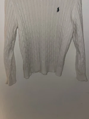 Vit cableknit från Polo Ralph Lauren - Kabelstickad vit tröja från Polo Ralph Lauren med klassisk rund hals och mörkblå broderad logga på bröstet. Tröjan har lång ärm och ribbade muddar. Perfekt för lager på lager under höst och vinter.