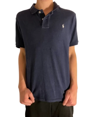 Mörkblå polo piké från Ralph Lauren - Säljer en mörkblå polo piké från Ralph Lauren med klassisk krage och två knappar framtill. T-shirten har korta ärmar och den ikoniska broderade loggan på bröstet. Materialet är mjuk bomull som känns skön mot huden.