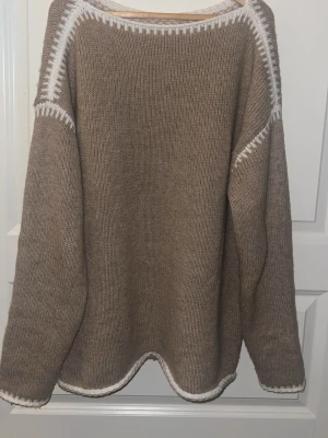 Beige stickad tröja med vit kant - Mysig beige stickad tröja med vit kant längs halsringning, axlar och nederkant. Tröjan har långärmade och en loose passform, perfekt för kyliga dagar. Enkel design med snygga detaljer som ger en chill vibe.