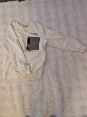 Vit sweatshirt från WOXO 720° - Vit sweatshirt från WOXO 720° med en stor grå ficka på bröstet och tryck i svart. Tröjan har rund halsringning, långa ärmar och ribbade muddar. Perfekt för dig som gillar en clean men ändå unik stil.