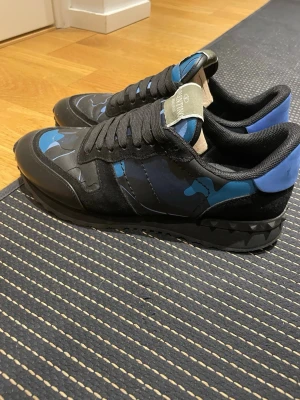 Valentino Garavani sneakers med blå camo - Snygga sneakers från Valentino Garavani med svart och blått camouflagemönster. Skorna har svarta snören, svarta och blå detaljer samt en cool nubbad sula. Materialmix av skinn och mocka ger en lyxig känsla. Perfekta för dig som vill sticka ut med stil.