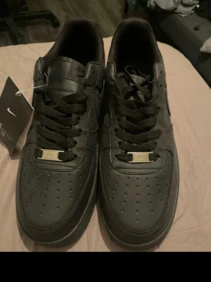 Nike Air Force 1 helsvarta sneakers - Säljer ett par klassiska Nike Air Force 1 i helsvart skinn. Skorna har svart sula, svarta skosnören och diskret Nike-logga på sidan och hälen. Perforerad tåbox för extra ventilation och metallbricka med 'AF1' på snörningen. Perfekta för dig som vill ha en clean och tidlös look.