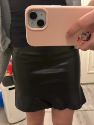 Svart kortkjol i skinnimitation - Säljer en svart kortkjol i skinnimitation med volangkant nedtill. Kjolen har en tight passform och ger en cool vibe till outfiten. Perfekt för dig som gillar trendiga detaljer och vill sticka ut lite extra.