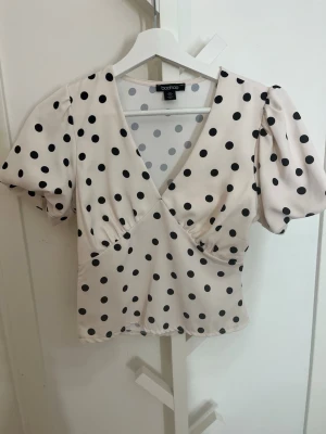 Prickig blus från Boohoo - Säljer en söt blus från Boohoo i ljusbeige med svarta prickar. Modellen har puffiga korta ärmar, v-ringning och figurnära passform. Perfekt för dig som gillar retrovibbar och vill sticka ut lite extra. Pris går att diskutera.