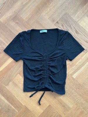 Svart ribbad topp med snörning - Säljer en svart ribbad topp. Toppen har korta ärmar, djup rundringning och snygg snörning framtill. Materialet är mjukt och stretchigt.