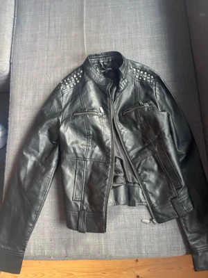 Svart skinnjacka med nitar New Look - Säljer en svart skinnjacka från New Look i storlek S. Jackan har coola silverfärgade nitar på axlarna, dragkedjor framtill och en klassisk bikerstil. Perfekt för dig som vill ha en edgy look och gillar detaljer som sticker ut.