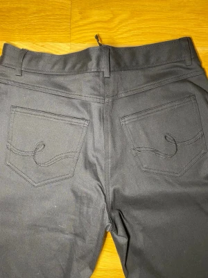 Svarta bootcut jeansbyxor - Säljer ett par svarta jeansbyxor med bootcut passform. Byxorna har klassiska fem fickor, bälteshällor och snygga broderade detaljer på bakfickorna. Materialet är slitstarkt och känns mjukt mot huden. Perfekta för dig som gillar en tidlös look.