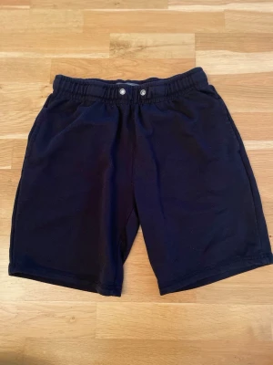 Svarta mjukisshorts med resårmidja - Säljer ett par svarta mjukisshorts med elastisk midja och två knappar framtill. Shortsen har en enkel design med en bakficka och är gjorda i mjukt bomullstyg. Perfekta för chill dagar eller träning.
