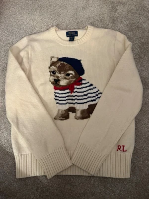 Vit stickad tröja Polo Ralph Lauren - Vit stickad tröja från Polo Ralph Lauren med ett stort motiv av en hund i randig tröja, blå basker och röd scarf på framsidan. RL-broderi i rött vid nederkanten. Rund hals och långa ärmar med ribbade muddar. Perfekt för dig som gillar unika och lekfulla plagg.