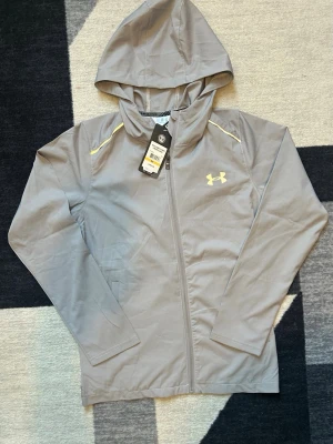 Grå vindjacka från Under Armour - Snygg grå vindjacka från Under Armour med huva och dragkedja. Jackan har diskreta gula detaljer vid axlarna och stort logotryck på ryggen. Tillverkad i lätt polyester, perfekt för träning eller när du vill ha en sportig look. Långärmad och med praktiska fickor framtill.