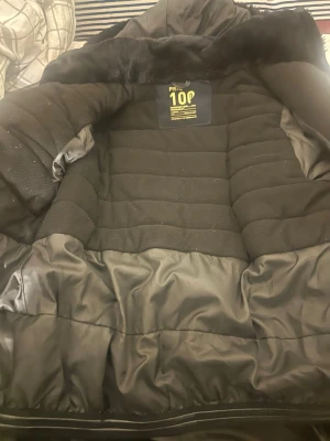 Svart pufferjacka med fuskpäls - Säljer en svart pufferjacka från Piste 100, designad i de franska alperna. Jackan har en stor, mjuk krage med fuskpäls och quiltade partier. Utsidan är i syntetmaterial och insidan är vadderad för extra värme. Perfekt för kalla dagar och riktigt snygg till vintern. M-L i storlek 