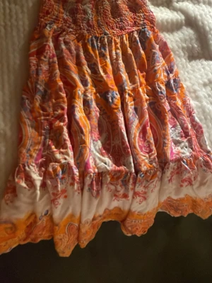 Färgglad kortkjol med paisleymönster - Säljer en superfin kortkjol med färgstarkt paisleymönster i orange, rosa och vitt. Kjolen har volanger och en smockad midja som ger en skön och avslappnad passform. Perfekt för varma dagar och ger en bohemisk vibe.