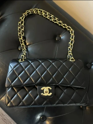 Svart väska - Ikonisk svart axelväska från…. skinn med klassisk guldfärgad kedja och CC-lås. Insidan är vinröd med präglad logga. Perfekt storlek för det viktigaste och en riktig statement piece för din outfit.