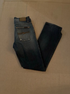 Nudie Jeans blå raka jeans - Säljer ett par klassiska blå jeans från Nudie Jeans med raka ben och snygga slitningar. Jeansen har fem fickor, orange sömmar och en mörk tvätt med ljusare partier. Tillverkade i bomull och har en normal passform. Perfekta för en avslappnad stil.