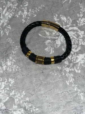 Ingen väst på bilden - Bilden visar ett svart armband med guldfärgade detaljer, inte en väst. Kan ej ge beskrivning eller taxonomier för väst.