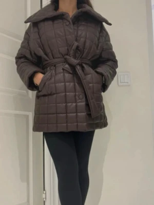 Brun quiltad pufferjacka med bälte - Säljer en brun quiltad pufferjacka med bred krage och knytbälte i midjan. Jackan har stora fickor och en oversized passform som ger en cool och avslappnad look. Perfekt för kyliga dagar när du vill vara både varm och snygg.