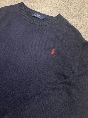 Mörkblå stickad tröja Polo Ralph Lauren - Mörkblå stickad tröja från Polo Ralph Lauren med klassisk rund hals och röd broderad logga på bröstet. Perfekt för lager-på-lager och passar till både jeans och chinos. Skön och stilren modell med lång ärm.