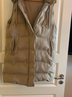Beige dunväst från Zara - Säljer en beige dunväst från Zara med huva och dragkedja framtill. Västen har två fickor med dragkedja och tryckknappar längs öppningen. Perfekt för lager på lager och riktigt snygg till höstlooken.