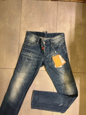 Blå Dsquared2 jeans med slitningar - Säljer ett par blå jeans från Dsquared2 med coola slitningar och färgstänk på benen. Jeansen har klassisk femficksmodell, knappgylf med röd detalj och tydliga tvättade partier för en trendig look. Perfekta för dig som gillar streetwear och vill sticka ut.