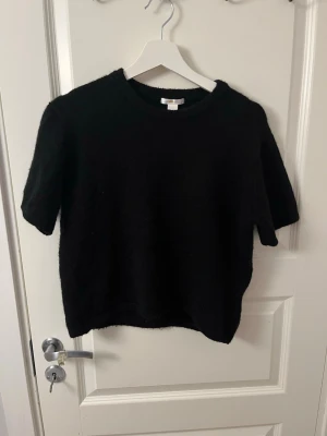 Svart stickad topp Gina Tricot - En svart stickad topp från Gina Tricot i storlek 170. Modellen har korta ärmar och rund halsringning. Materialet är mjukt och lite luddigt, vilket ger en mysig känsla. Perfekt att styla med jeans eller kjol för en enkel men snygg look.