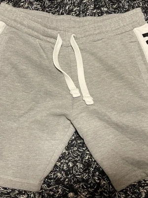 Grå Arsenal shorts med snörning Storlek Small - Snygga grå shorts från Arsenal med vit snörning i midjan och två bakfickor. Mjukt material i bomull och polyester, perfekt för chill eller sport. Detalj med 'Gunners 1886' på ena bakfickan och klubbmärke på insidan.