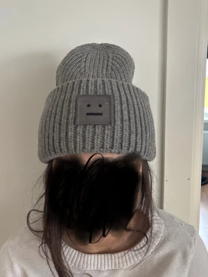 Grå stickad mössa från Acne Studios - Säljer en grå ribbstickad mössa från Acne Studios med deras ikoniska ansiktslogga framtill. Mössan är i mjuk ull och har en bred uppvikt kant för extra stilpoäng. Perfekt accessoar för att hålla värmen och lyfta din outfit.