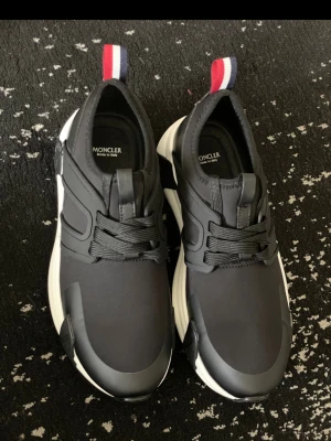 Svarta sneakers från Moncler - Säljer ett par svarta Moncler sneakers med vita sulor och svarta snören. Skorna har en sportig design med detaljer i syntet och textil samt Monclers klassiska röd-vit-blå flärp bak. Perfekta för dig som gillar stilrena och exklusiva sneakers. Storlek 44 passar 43 med 