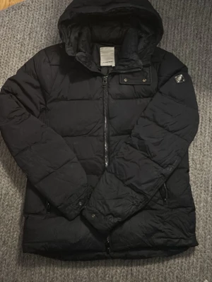 Dunjacka J.Lindeberg - Säljer en svart pufferjacka från J.Lindeberg. Jackan har en stilren look med diskret logga på ärmen. Perfekt för kalla dagar och riktigt skön att bära. 