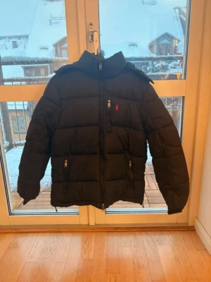 Svart pufferjacka från Polo Ralph Lauren - Säljer en svart pufferjacka från Polo Ralph Lauren med hög krage och huva. Jackan har dragkedja framtill, två sidofickor med dragkedja och en liten röd logga på bröstet. Materialet är polyester och jackan har långa ärmar med resår vid handlederna.