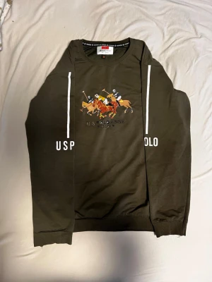 Grön sweatshirt från US Polo Assn - Mörkgrön sweatshirt från US Polo Assn med stort tryck av polospelare på bröstet och vita detaljer längs ärmarna. Tröjan har rund halsringning och ribbade muddar vid ärmslut och nederkant. Perfekt för en sportig och avslappnad stil.