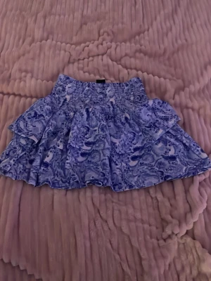 Blåvit volangkjol med mönster - Supersöt kort kjol i blått och vitt med volanger och elastisk midja. Kjolen har ett unikt abstrakt mönster och är perfekt för dig som vill ha en lekfull och trendig look. Lätt och luftig, passar perfekt till varma dagar.