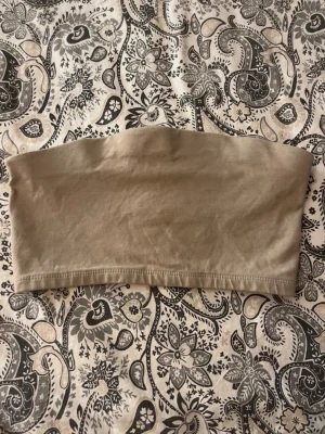 Beige bandeau-topp – aldrig använd - Beige bandeau-topp i mjukt och stretchigt material. Enkel och stilren modell som passar perfekt till både vardag och sommaroutfits. Toppen är aldrig använd och i nyskick. Lätt att matcha med jeans, kjol eller under kavaj.