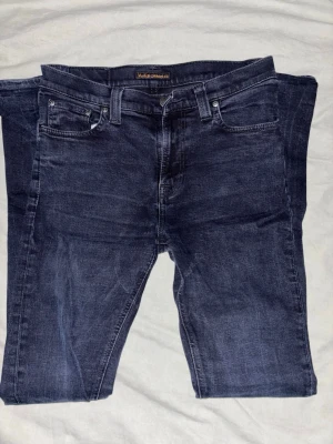 Mörkblå jeans från Nudie Jeans - Snygga mörkblå jeans från Nudie Jeans med klassisk femficksdesign och subtila sömmar på bakfickorna. Jeansen har rak passform och är tillverkade i mjukt bomullsmaterial med en lätt tvättad look. Perfekt för dig som gillar stilrena och tidlösa jeans.