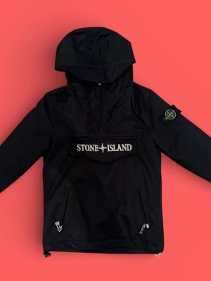 Svart vindjacka från Stone Island - Säljer en svart vindjacka från Stone Island med huva och stor logga på bröstet. Jackan har dragkedja framtill, två sidofickor med dragkedja och klassisk patch på ärmen. Tillverkad i lätt polyester och har justerbar nederkant.