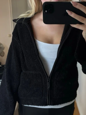 Svart teddyjacka med dragkedja - Mysig svart teddyjacka med hel dragkedja framtill. Jackan har två stora fickor och är tillverkad i fluffigt fleece-material. Perfekt för lager på lager under höst och vinter.