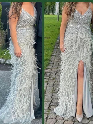 Festklänning  - Säljer en galet snygg silverfärgad långklänning med v-ringning och glittriga paljettdetaljer upptill. Kjolen är täckt av dekorativa fjädrar och har en hög slits framtill. Den är köpt från SE fashion och har bara använts i några timmar🫶🏼M-L