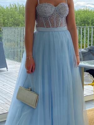 Ljusblå långklänning med glittrig topp - Säljer en ljusblå långklänning med glittrig, pärlbeströdd topp och smala axelband. Kjolen är i flera lager av tunn tyll som ger ett svävande intryck. Toppen är som en korsett med transparenta detaljer och gnistrande pärlor. Perfekt för bal eller student.