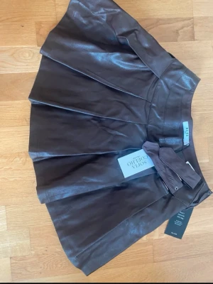 Plisserad brun skinnkjol från NA-KD - Säljer en brun plisserad kortkjol i skinnimitation från NA-KD x Sofia Coelho. Kjolen har bred midja med dubbla bältesdetaljer och dragkedja i sidan. Snygg och edgy modell som ger en cool vibe till outfiten. Helt ny och aldrig använt. Väldigt söt och har en annan det är därför jag säljer ena.