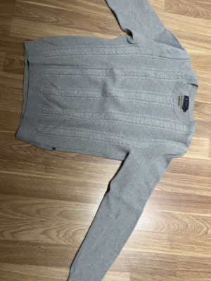 Grå kabelstickad tröja från Riley - Snygg grå kabelstickad tröja från Riley i premium bomull. Tröjan har rund hals, ribbade muddar och lång ärm. Perfekt för lager-på-lager och passar till både jeans och kjol. Klassisk och stilren design som funkar året om.